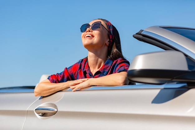 Vrolijke aziatische vrouw met een zonnebril die in een cabrio zit en glimlacht en omhoog kijkt