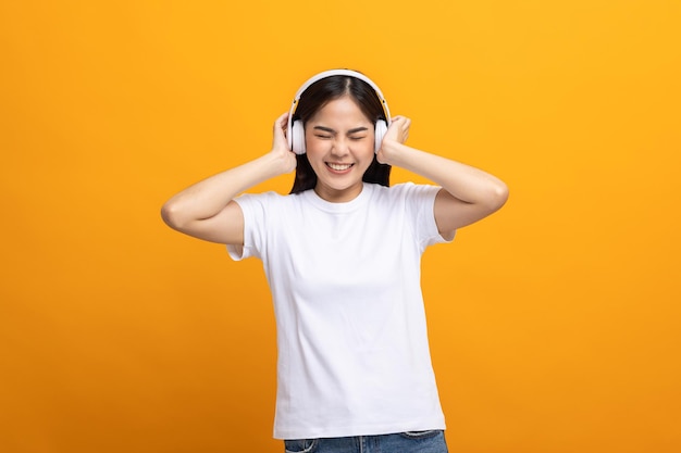 Vrolijke Aziatische vrouwelijke tiener luistert naar de muziek met witte koptelefoon dansen op geïsoleerde gele achtergrond. Mooie jonge vrouw in de hand raakt een draadloze hoofdtelefoon aan die plezier heeft met muziek.