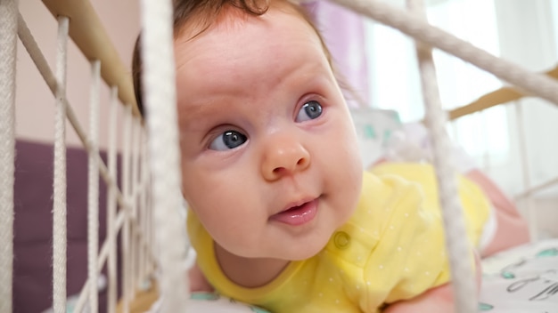 Vrolijke babymeisje in gele bodysuit ligt op buik in schommelwieg spelen en kijken met belangstelling tegen raam in kamer extreme close-up