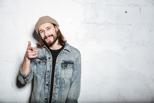 Foto vrolijke bebaarde hipster man met hoed gekleed in spijkerjasje poseren over muur en wijzend