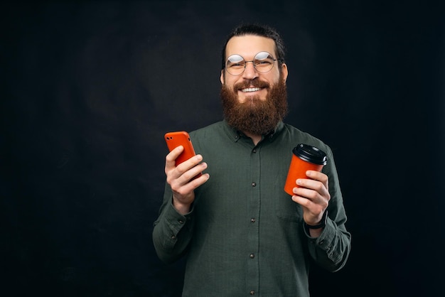 Vrolijke bebaarde hipster man met smartphone en koffie om over donkere achtergrond te gaan en naar de camera te kijken