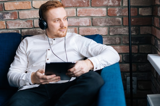 Vrolijke bebaarde jongeman in koptelefoon zittend op blauwe zachte bank in de woonkamer met zijn tabletcomputer en weg kijken. Concept van muziek ontspanning.