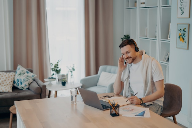 Vrolijke bebaarde jongeman met koptelefoon met online vergadering die videogesprek voert op laptop terwijl hij op een gezellige werkplek aan huis werkt, zittend in een moderne woonkamer Remote job concept