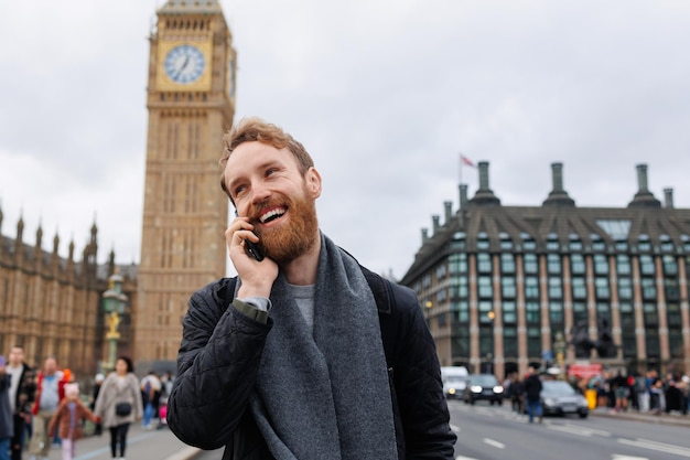 Vrolijke bebaarde man die aan de telefoon praat terwijl hij op de achtergrond van de Big Ben-toren staat
