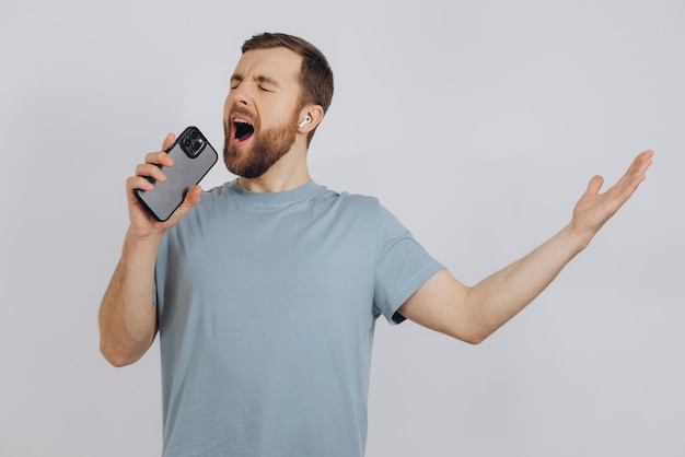 Vrolijke, bebaarde man die danst met een koptelefoon, luisterend naar muziek in een koptelefoon met een mobiele telefoon en glimlachend blij dat hij over een witte achtergrond staat