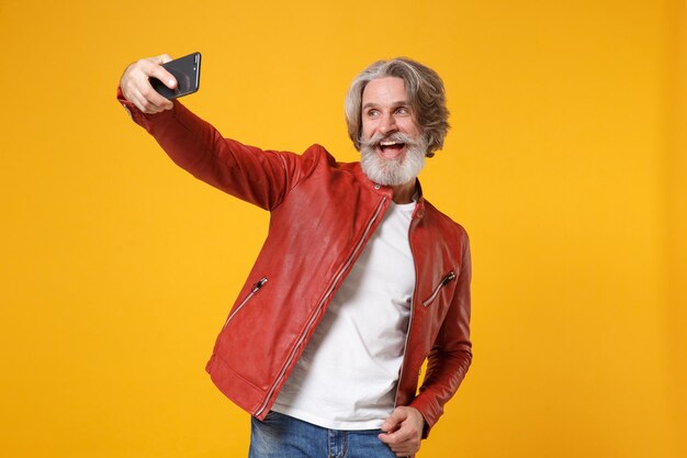 Vrolijke bejaarde grijsharige snor bebaarde man in rood leren jas poseren geïsoleerd op geeloranje achtergrond. Mensen levensstijl concept. Mock-up kopie ruimte. Selfie geschoten op mobiele telefoon.