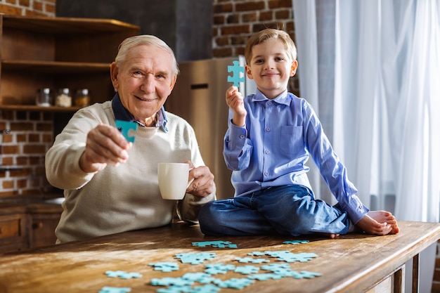 Vrolijke bejaarde man en zijn kleine schattige kleinzoon die puzzels in de keuken samenstellen