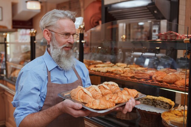 Foto vrolijke bejaarde mannelijke bakker die croissants draagt om bij zijn bakkerijwinkel te bakken