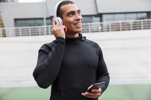 Vrolijke blanke sportman in trainingspak met koptelefoon en mobiele telefoon tijdens het trainen in het stadion na regen