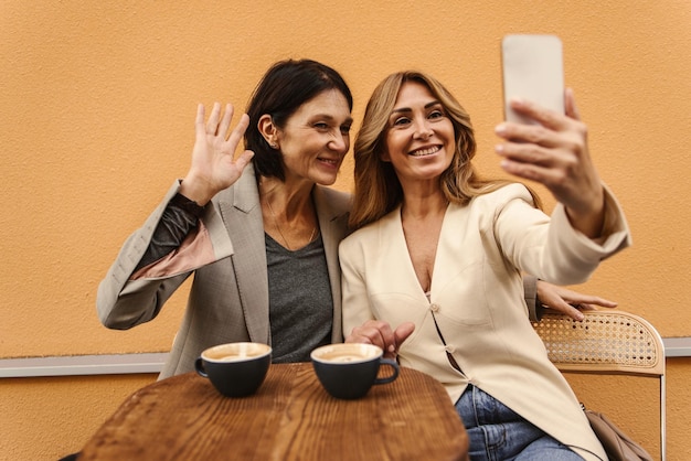 Vrolijke blanke vrouwen van middelbare leeftijd die samen foto's maken op smartphone terwijl ze buiten koffie drinken Vrije tijd voor selfies