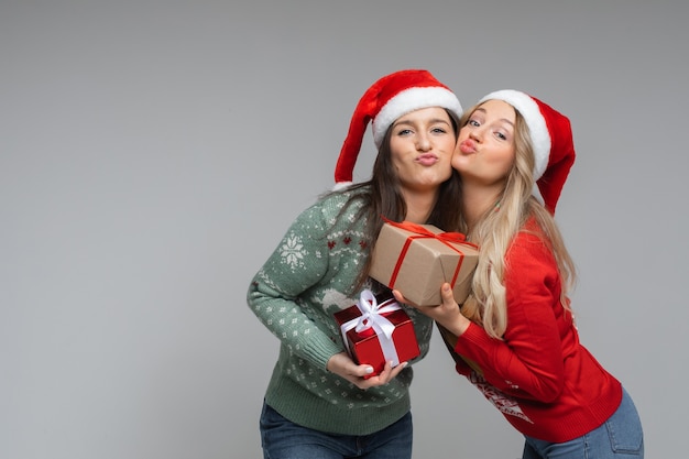 Vrolijke blanke zusters in warme groene en rode truien houden hun kerstcadeaus vast, foto geïsoleerd op een witte muur