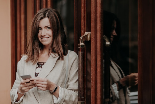 Vrolijke blije vrouw met donker steil haar gebruikt smartphone gekleed in elegante kleding kijkt positief naar camera geniet van online communicatie controleert route stuurt berichten leest melding