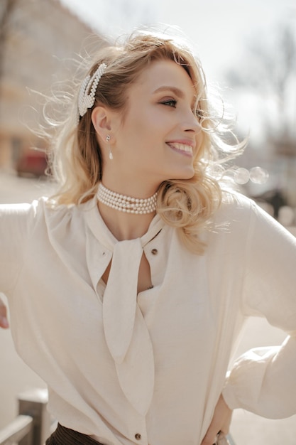 Vrolijke blonde krullende jonge vrouw in stijlvolle parelketting en witte trendy blouse glimlacht oprecht en loopt naar buiten
