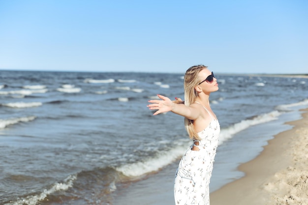 Vrolijke blonde mooie vrouw op het oceaanstrand in een witte zomerjurk en zonnebril, open armen.