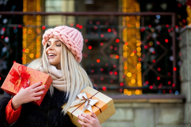 Vrolijke blonde vrouw gekleed in warme outfit, met geschenkdozen op de achtergrond van bokeh lichten. Ruimte voor tekst