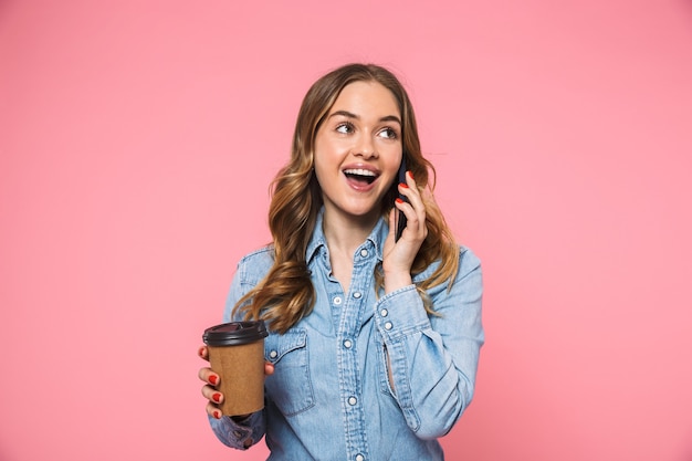 Vrolijke blonde vrouw in denim shirt die koffie drinkt en praat met een smartphone terwijl ze wegkijkt