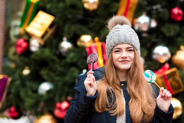 Vrolijke blonde vrouw in warme muts die plezier heeft met lolly op de sparren achtergrond