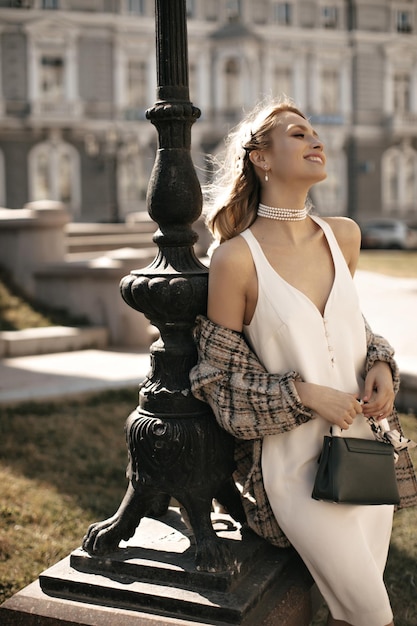 Vrolijke blonde vrouw in zijde witte elegante jurk parelketting en geruit jasje leunt op straatlantaarn en houdt handtas charmante dame poses op stadsplein