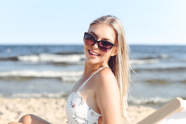 Vrolijke blonde vrouw met een zonnebril en ontspannend op een houten ligstoel aan het oceaanstrand