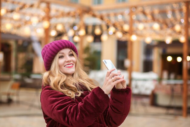 Vrolijke blonde vrouw selfie te nemen op de mobiele telefoon op de achtergrond van kerstverlichting