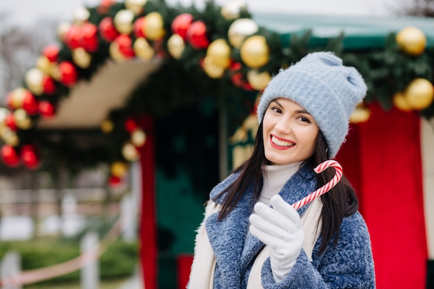 Vrolijke brunette vrouw draagt gebreide muts en winterjas met snoep in de buurt van de straat kerstversiering met ballen. Ruimte voor tekst