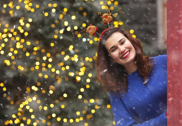 Vrolijke brunette vrouw draagt grappige haarhoepel die plezier heeft bij de kerstboom tijdens sneeuwval. Lege ruimte