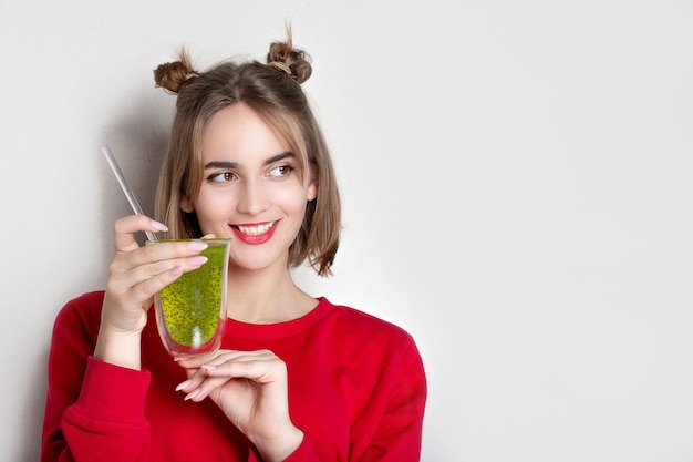 Vrolijke brunette vrouw draagt rode trui met glas groen sap met chiazaad over een grijze muur. Lege ruimte