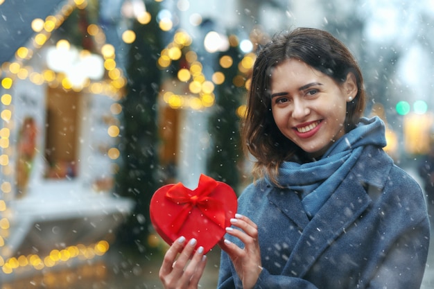 Vrolijke brunette vrouw met geschenkdoos in de buurt van de kerstboom tijdens de sneeuwval. Ruimte voor tekst