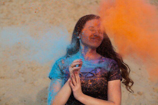 Vrolijke brunette vrouw met lang krullend haar die speelt met exploderend oranje en blauw droog Holi-poeder