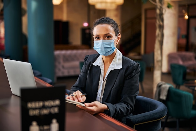 Vrolijke brunette vrouwelijke persoon die een steriel masker draagt tijdens het werken in een openbare ruimte