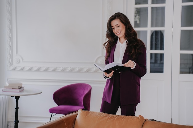 Vrolijke brunette zakenvrouw die een notitieboekje vasthoudt en haar week plant in een violet pak