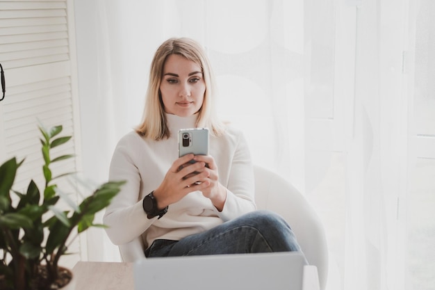 Vrolijke charmante vrouw die laptop en mobiele telefoon gebruikt die glimlacht terwijl ze op de werkplek zit