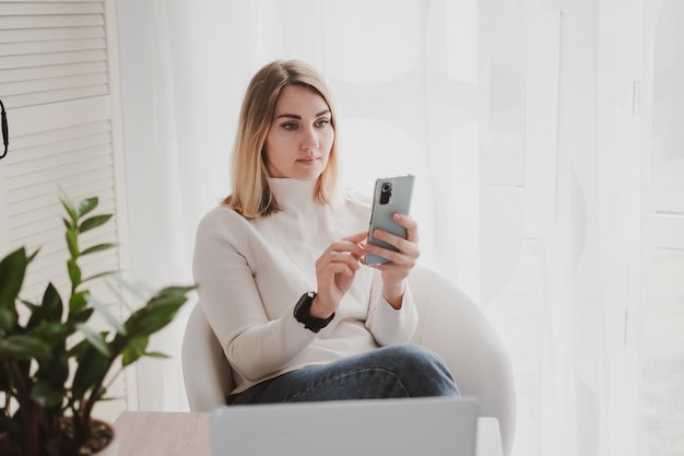 Vrolijke charmante vrouw die laptop en mobiele telefoon gebruikt die glimlacht terwijl ze op de werkplek zit