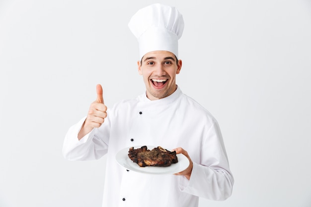 Vrolijke chef-kok die eenvormige paprika gekookte maaltijd van het biefstuklapje draagt op een plaat die over witte muur wordt geïsoleerd
