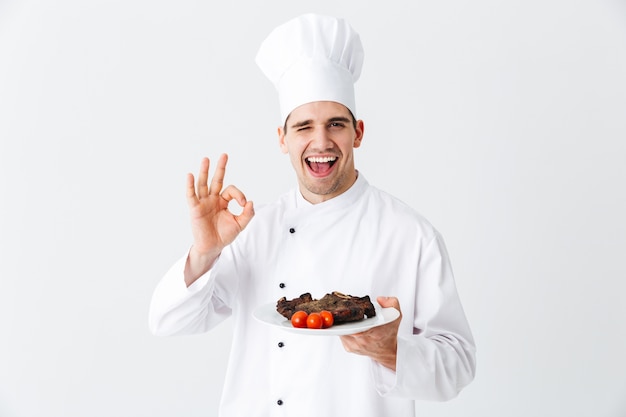 Vrolijke chef-kok die eenvormige paprika gekookte maaltijd van het biefstuklapje draagt op een plaat die over witte muur wordt geïsoleerd