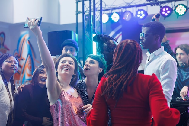 Vrolijke clubbers maken groepsfoto op smartphone terwijl ze feesten op een drukke dansvloer van een nachtclub met lichtjes. Zorgeloze glimlachende vrouw die mobiele telefoon selfie maakt met vrienden in de club