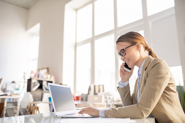 Vrolijke communicatieve jonge zakenvrouw met paardenstaart zittend aan een bureau en met behulp van laptop tijdens het praten over mobiel en werken met de klant