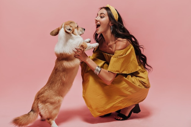 Vrolijke dame met lang donkerbruin haar in bandana moderne jurk en zwarte hakken spelen met kleine hond op geïsoleerde achtergrond