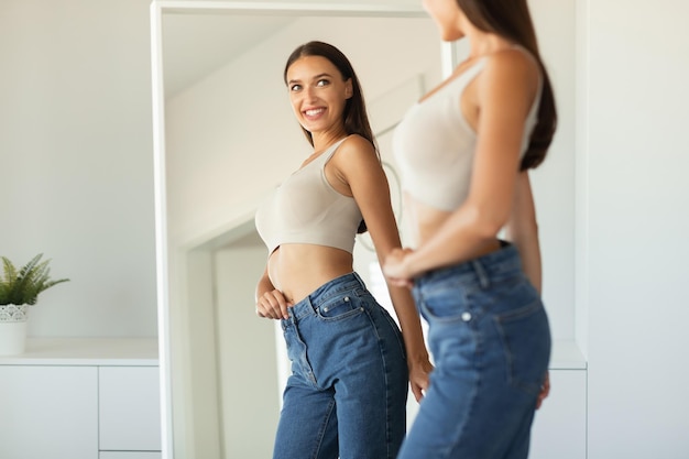 Foto vrolijke dame poseren met skinny jeans in de buurt van spiegel die binnenshuis staat