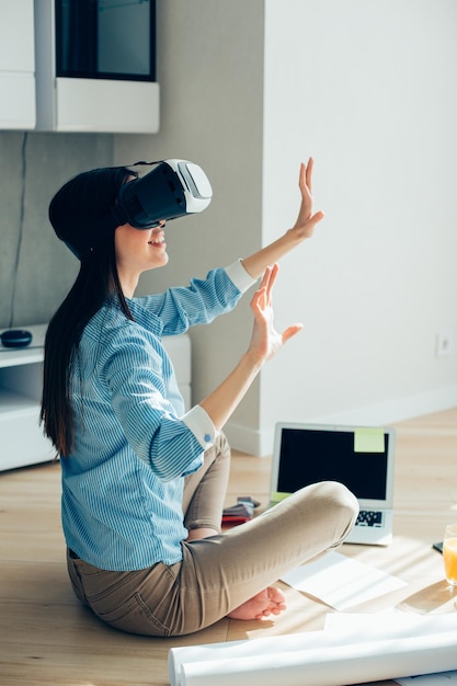 Vrolijke dame zit in kleermakerszit op de grond voor een laptop en draagt een virtual reality-apparaat