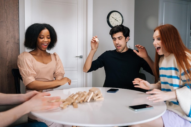 Vrolijke diverse mannen en vrouwen spelen met houten torenstukken op tafel en hebben plezier met vierkante blokken en houten kubussen die vallende houten toren vangen