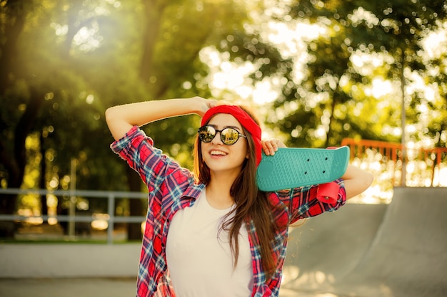 Vrolijke emotionele jonge vrouw gekleed in stijlvolle kleding terwijl ze een skateboard in haar hand houdt in het skatepark op een zonnige dag