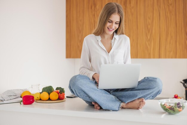 Vrolijke en lachende blonde vrouw die bovenop de tafel in de keuken zit en recepten zoekt op internet op de laptop