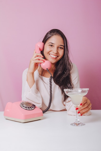 Vrolijke en lachende jonge brunette vrouw met drankje, maakt gebruik van een vintage telefoon. Portret op een roze ondergrond