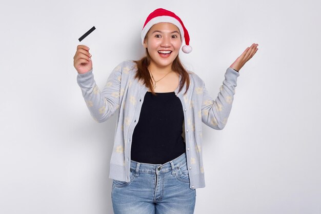 Vrolijke en opgewonden jonge Aziatische vrouw in een kerstmuts met een lege witte creditcard op camera geïsoleerd op een witte achtergrond