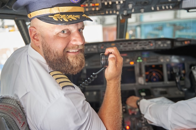 Foto vrolijke ervaren vlieger met een baard wendt zich tot de camera in een cockpit met een draagbare radiozender in de hand