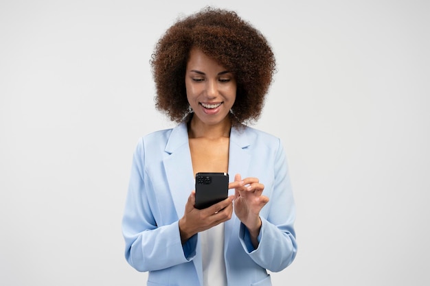 Vrolijke etnische vrouw met afrohaar die smartphone gebruikt die sociale inhoud controleert op witte achtergrond