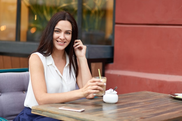 Vrolijke Europese vrouw geniet van vrije tijd, drinkt milkshake in openluchtcafetaria, brengt weekend in de grote stad door, wacht op telefoontje van smartphone, ziet er positief uit, gekleed in witte stijlvolle blouse