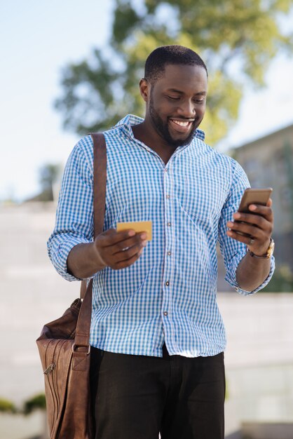 Vrolijke expressieve aantrekkelijke man die op straat staat en een bestelling plaatst met behulp van een speciale app en ervoor betaalt met zijn creditcard