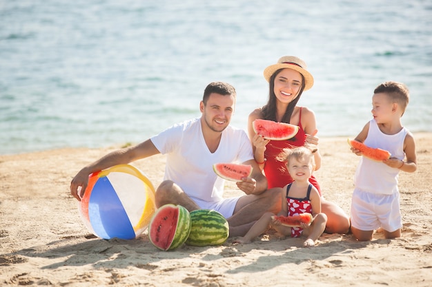 Vrolijke familie die watermeloen op het strand eet. Kleine kinderen en hun ouders aan de kust met plezier. Vrolijke familie aan de kust